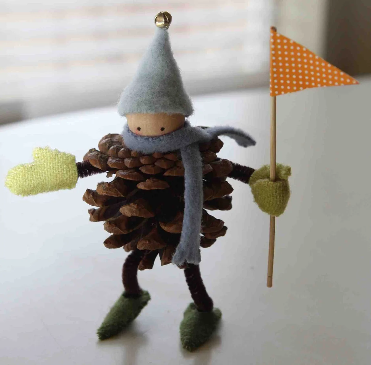 A handmade figurine crafted from a pinecone, featuring a felt hat with a bell, a gray scarf, green mittens, and green felt…