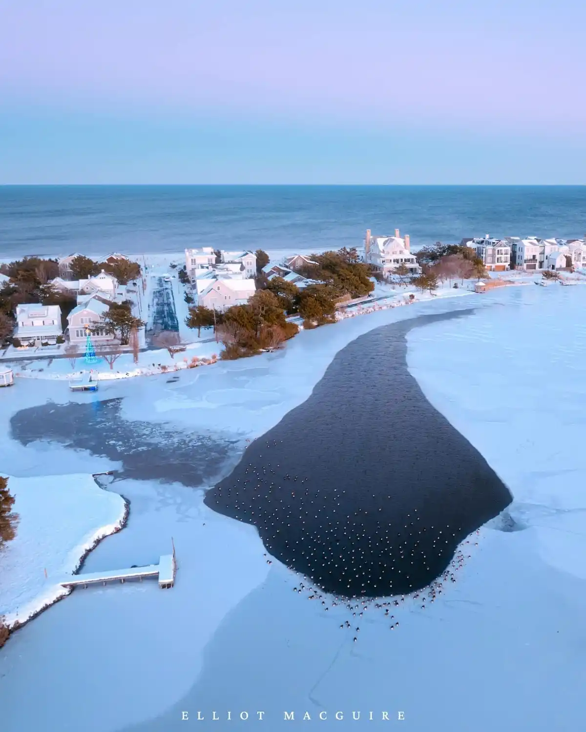 An aerial photo shows a frozen coastal area with snow-covered houses and a large, dark, unfrozen channel of water cutting…