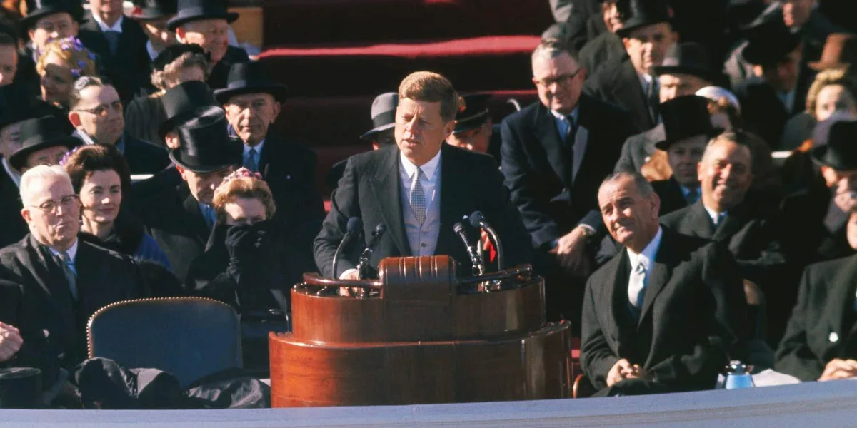 A historical photo shows John F. Kennedy speaking at a podium during his inauguration, with Lyndon B. Johnson seated beside…