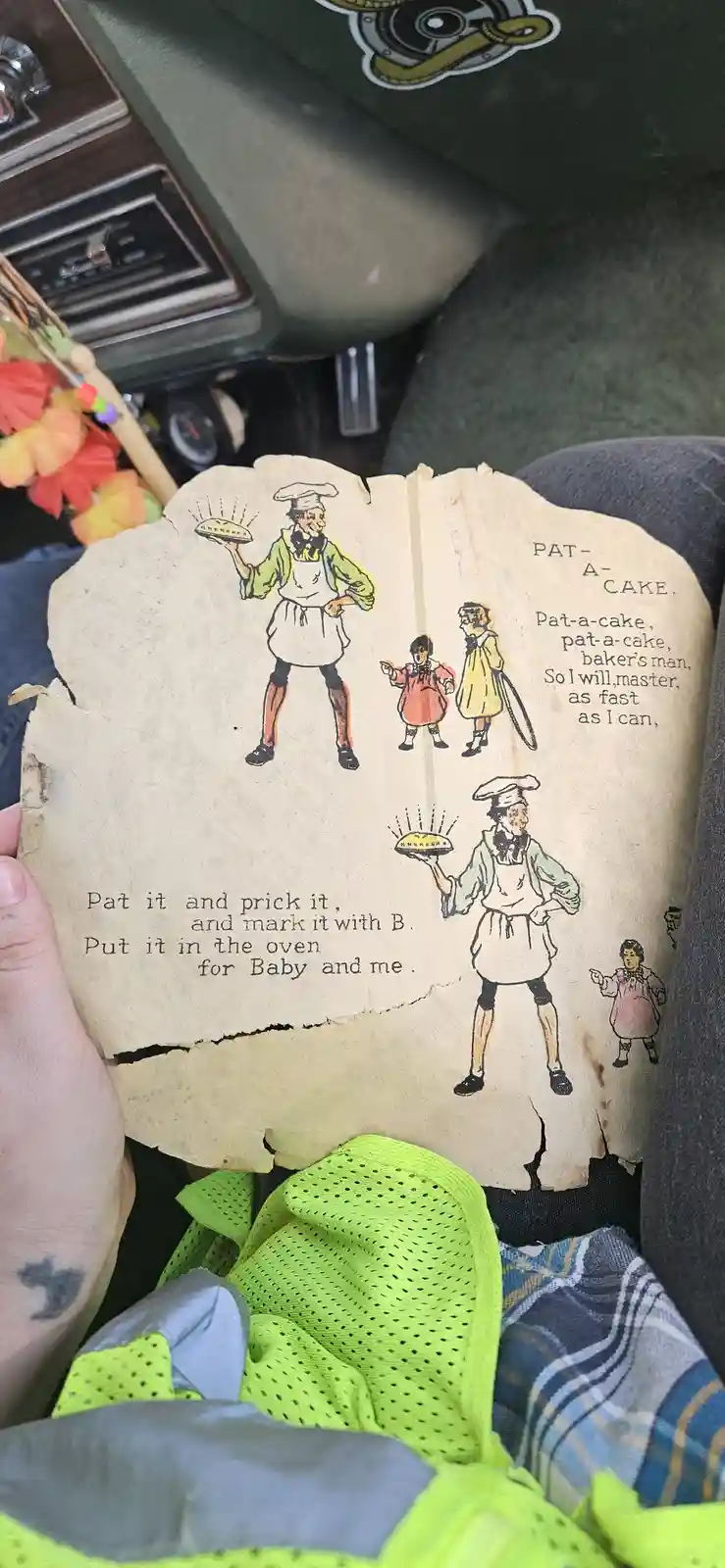 A person holds up a torn, vintage children's book page showing illustrations of a baker and children performing the…