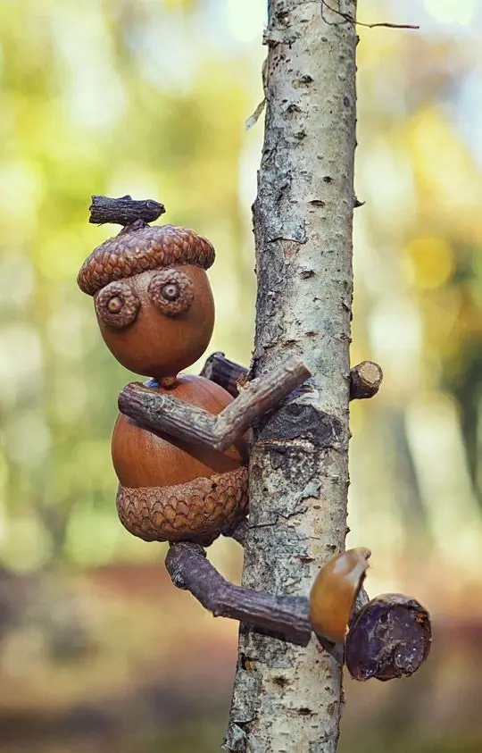 A whimsical woodland creature crafted from acorns and twigs clings to a tree trunk, with its acorn cap serving as a hat and…