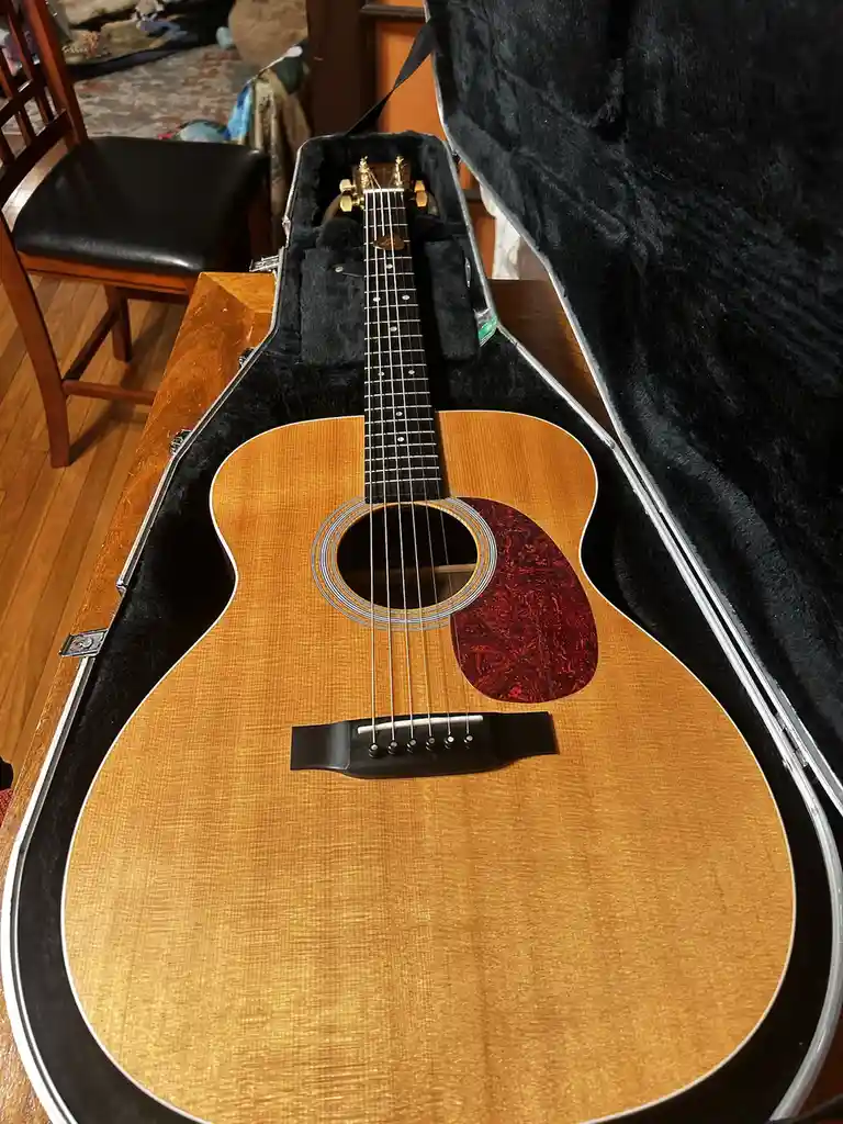 An acoustic guitar with a light wood finish and red purfling is displayed inside an open, black-lined hard case resting on a…