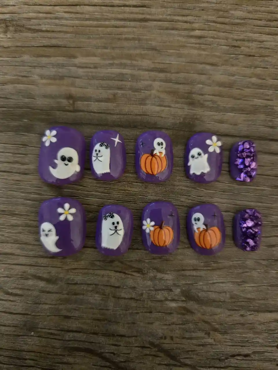 Ten purple Halloween-themed nail art pieces are arranged in two rows on a wooden surface. Each nail features cute cartoon…