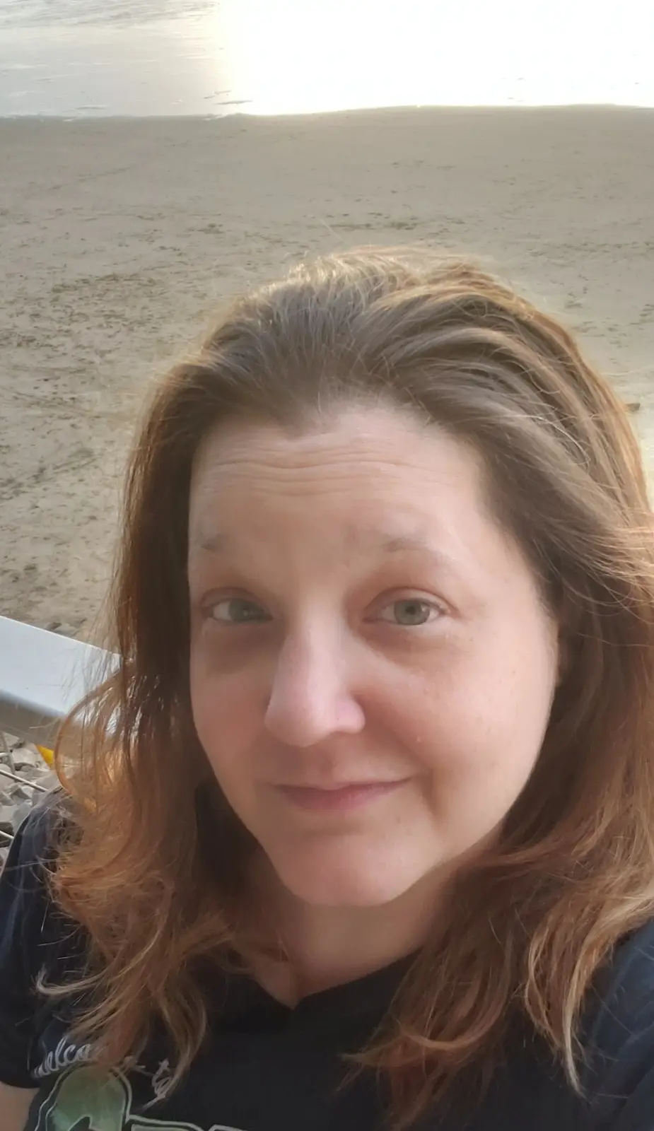 A close-up selfie of a woman with shoulder-length brown hair, smiling slightly while sitting on a beach with sand and water…
