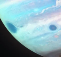 A close-up of Jupiter's swirling atmosphere showing two distinct dark oval-shaped spots, resembling eyes, set against…