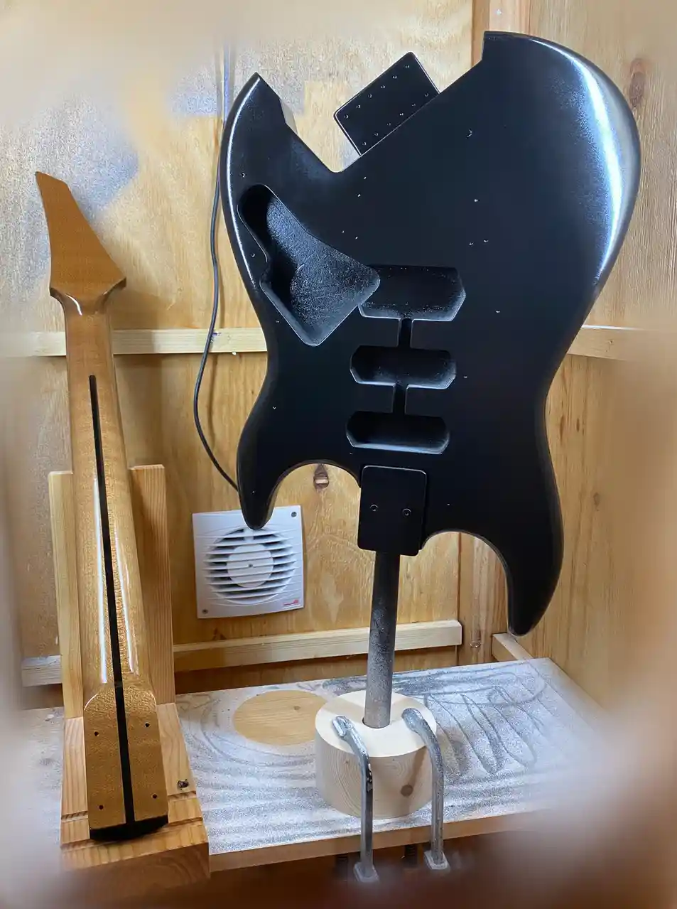A black, glossy electric guitar body is mounted vertically on a wooden stand in a workshop, next to an unfinished light wood…