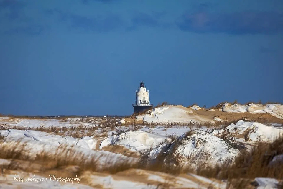 A white lighthouse stands in the distance atop sandy dunes covered in patches of snow under a blue sky. The foreground shows…