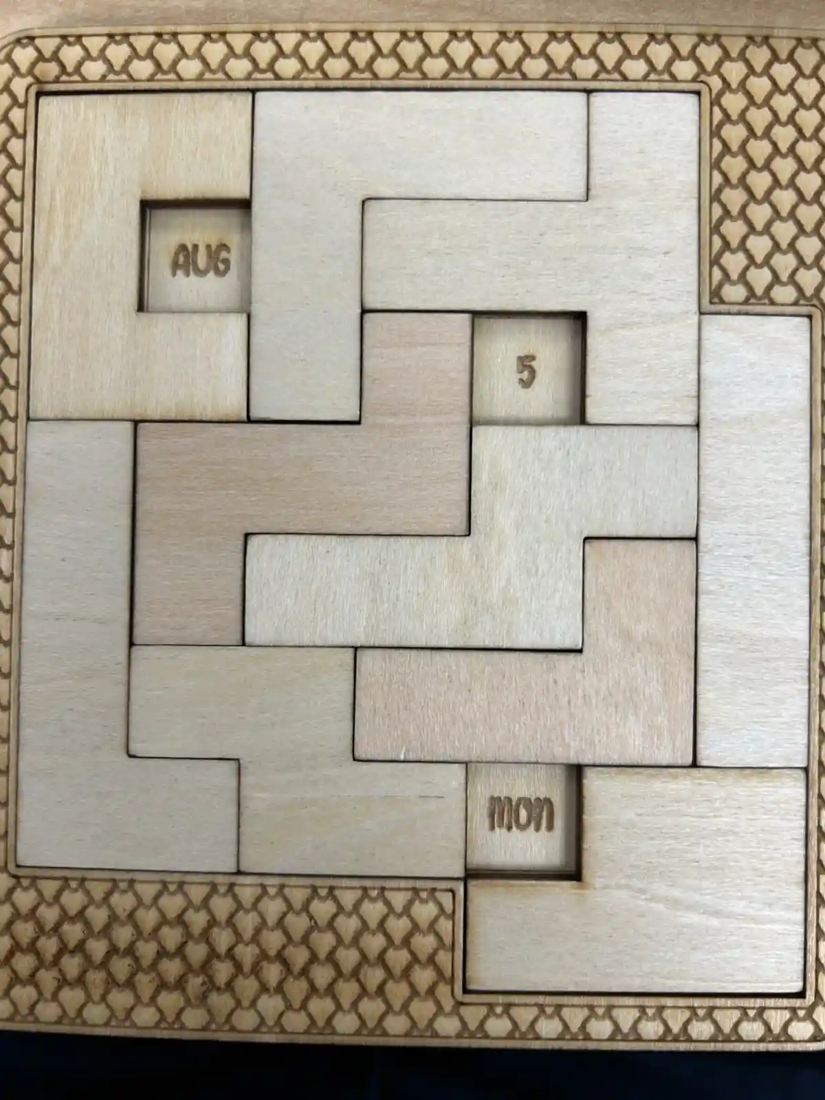 A wooden puzzle with interlocking pieces forming a grid, featuring engraved text in three recessed squares: 'AUG' in the…