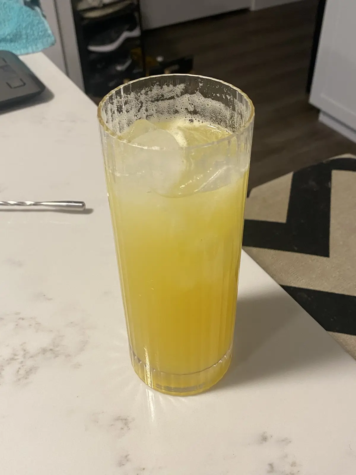 A tall, fluted glass filled with a yellowish beverage and ice sits on a white marble countertop, with foam residue around…