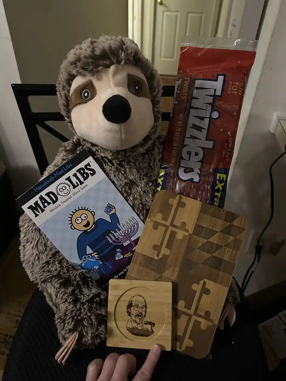 A plush sloth sits on a chair holding a 'Hanukkah Mad Libs' book, a bag of Twizzlers, and two wooden coasters—one with a…