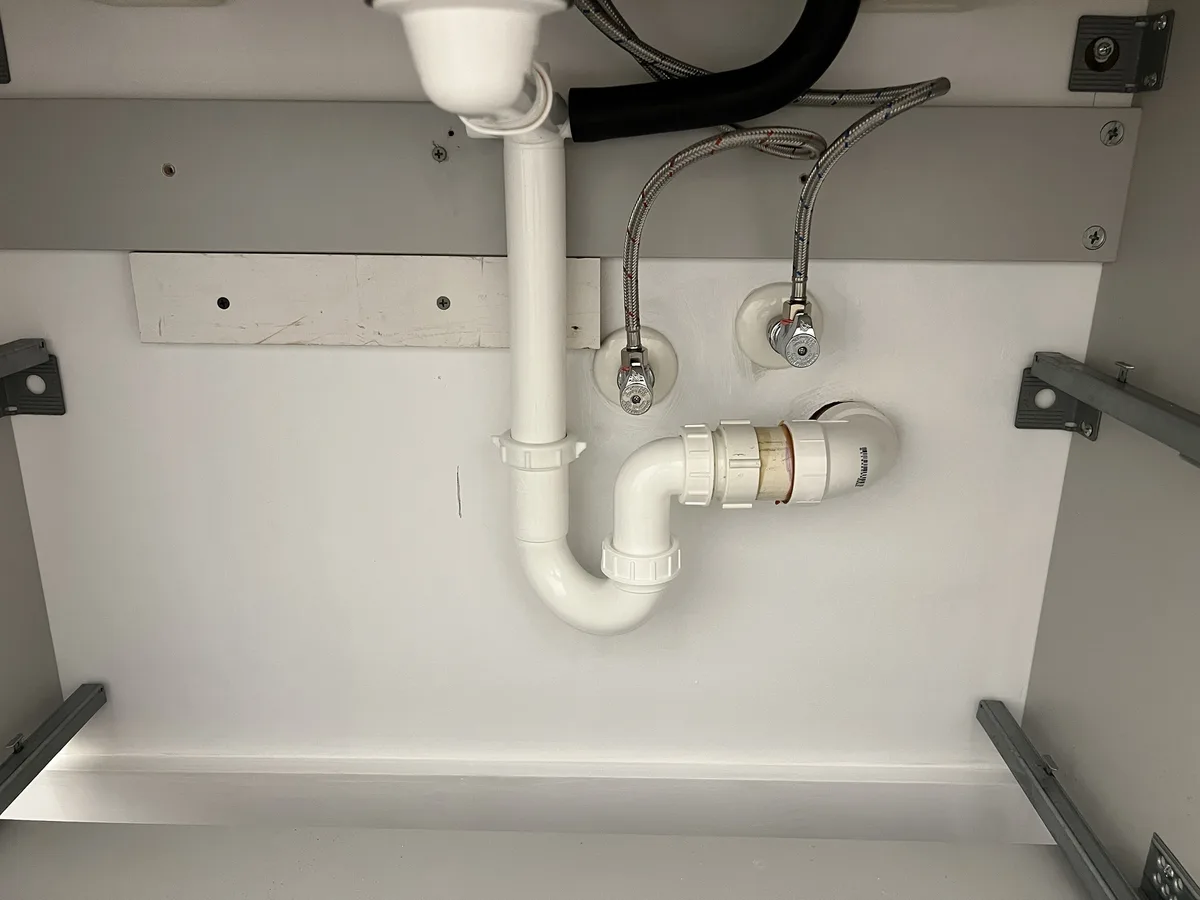 A close-up photo shows plumbing under a sink, featuring white PVC pipes, two chrome faucets with braided hoses, and a white…