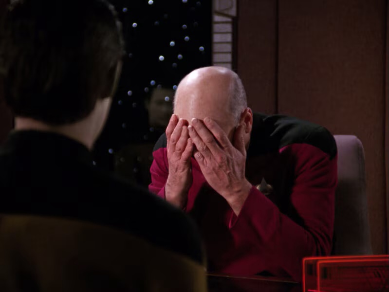 A bald man in a red Starfleet uniform sits at a table, covering his face with both hands in apparent distress or…