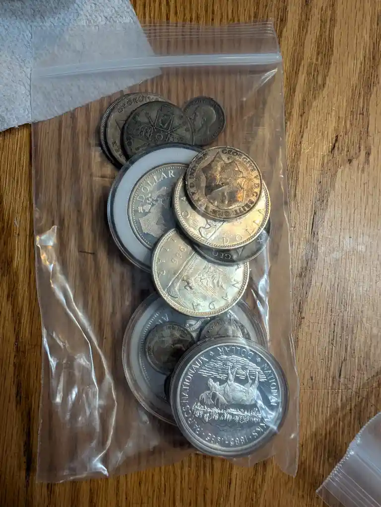 A clear plastic zip-top bag on a wooden surface contains a collection of various old coins, some in protective circular…