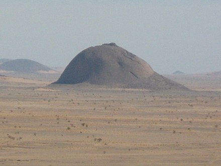 a wide shot of a large, smooth, dome-shaped rock formation in a barren, arid landscape with distant hills under a hazy sky…