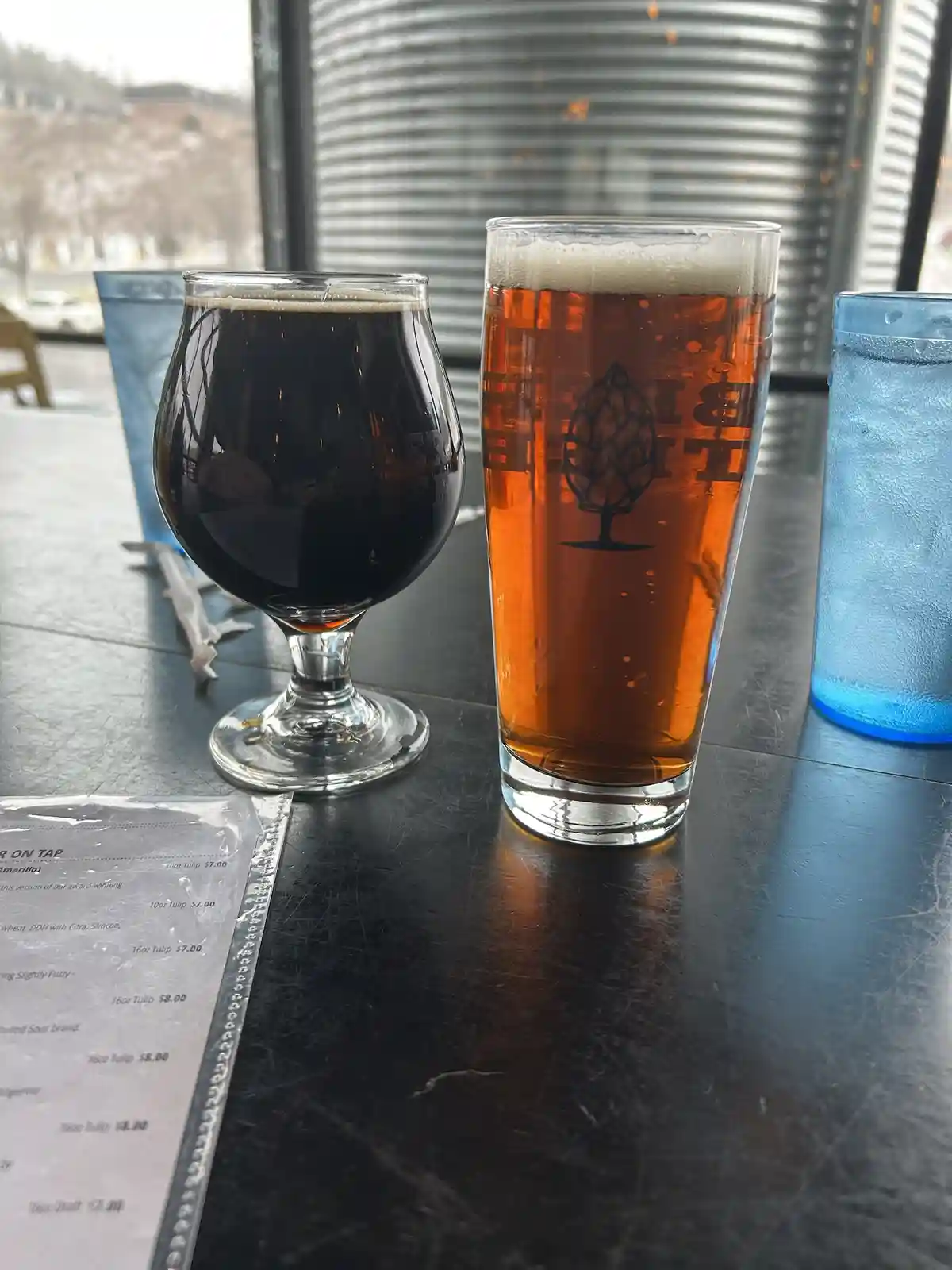 Two glasses of beer sit on a dark table: a dark stout in a tulip glass and a lighter amber ale in a pint glass with a hop…