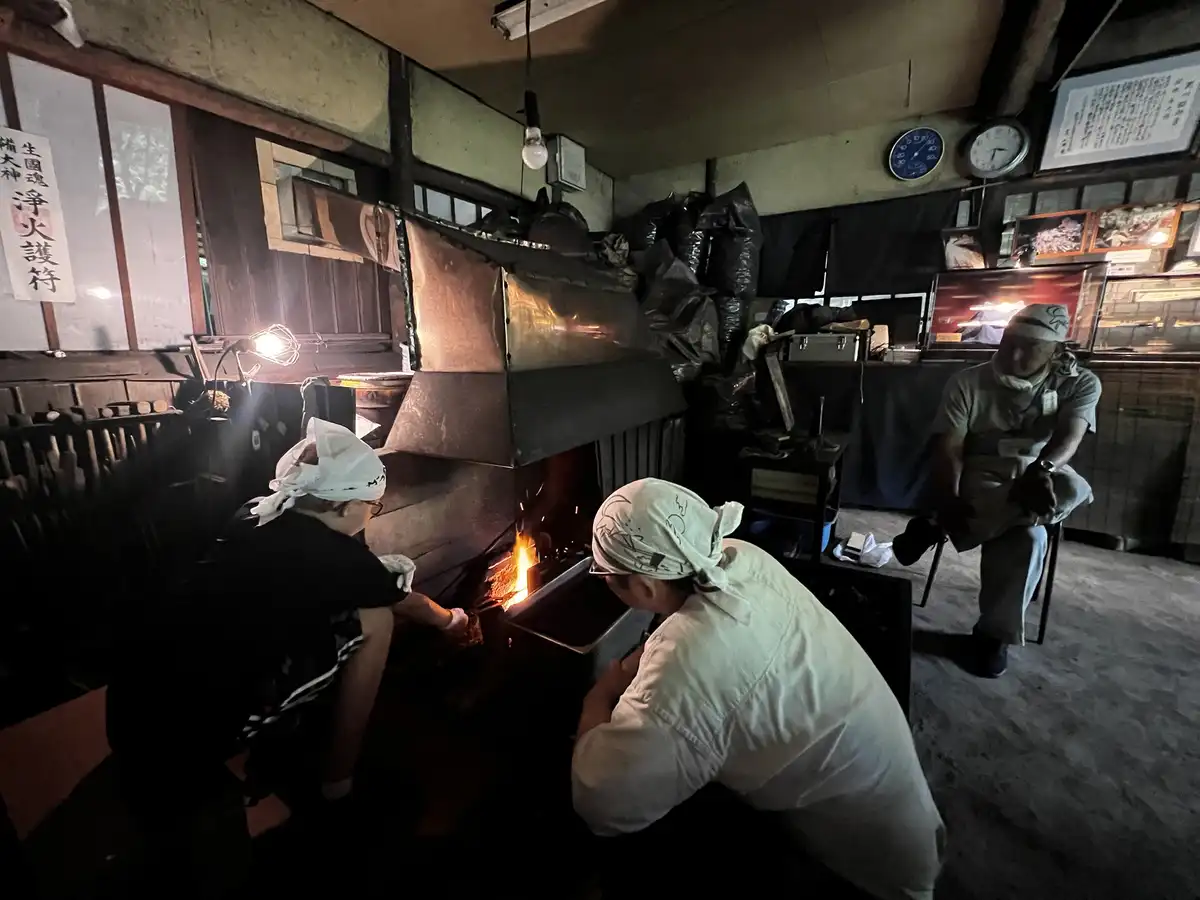 In a dimly lit, rustic Japanese forge, two workers in headscarves lean over a glowing furnace, tending to the fire while a…