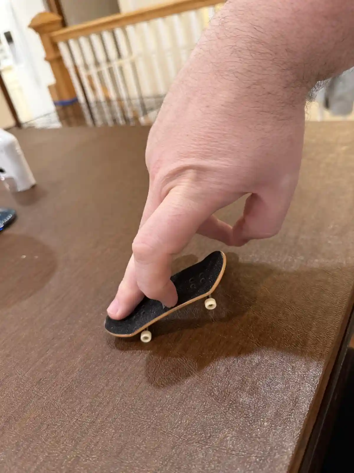 A person's hand is holding a tiny fingerboard skateboard on a brown textured table, with a wooden staircase railing visible…