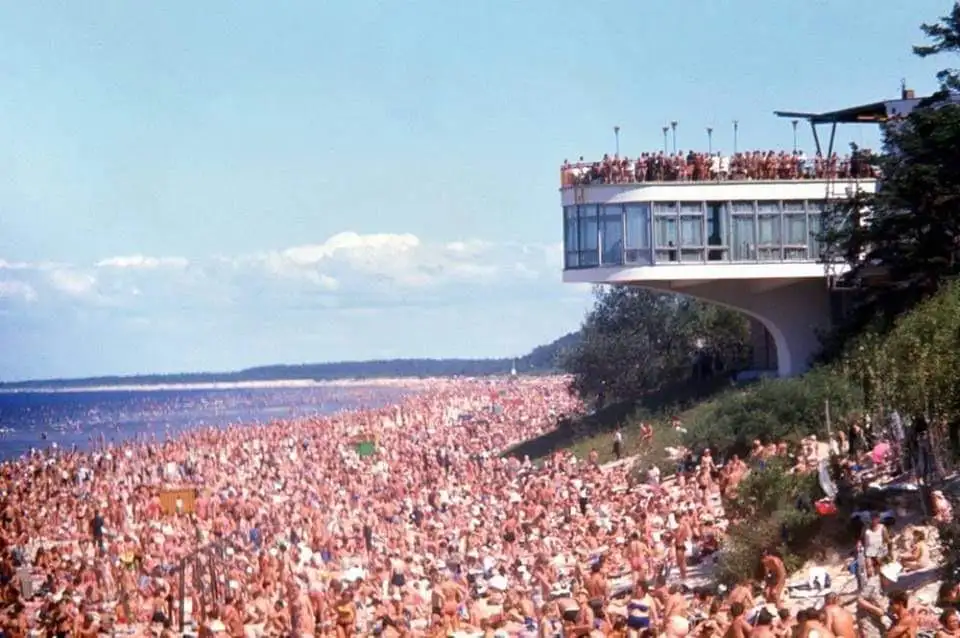 A vintage photo shows a densely packed beach with thousands of sunbathers, many in swimwear, stretching from the shoreline…