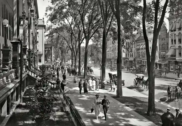 A vintage black-and-white photograph shows a bustling early 20th-century city street with pedestrians, horse-drawn…