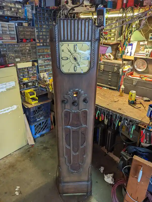 A tall, dark wooden Art Deco-style clock stands in a cluttered electronics workshop filled with tools, wires, and component…