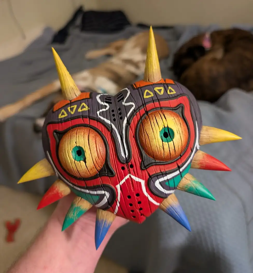 A hand holds a colorful, hand-carved wooden replica of Majora's Mask from The Legend of Zelda, featuring red, yellow, and…