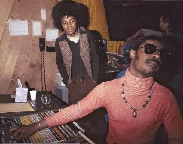 A vintage photo shows two Black men in a recording studio. In the foreground, Sly Stone, wearing sunglasses, a pink…