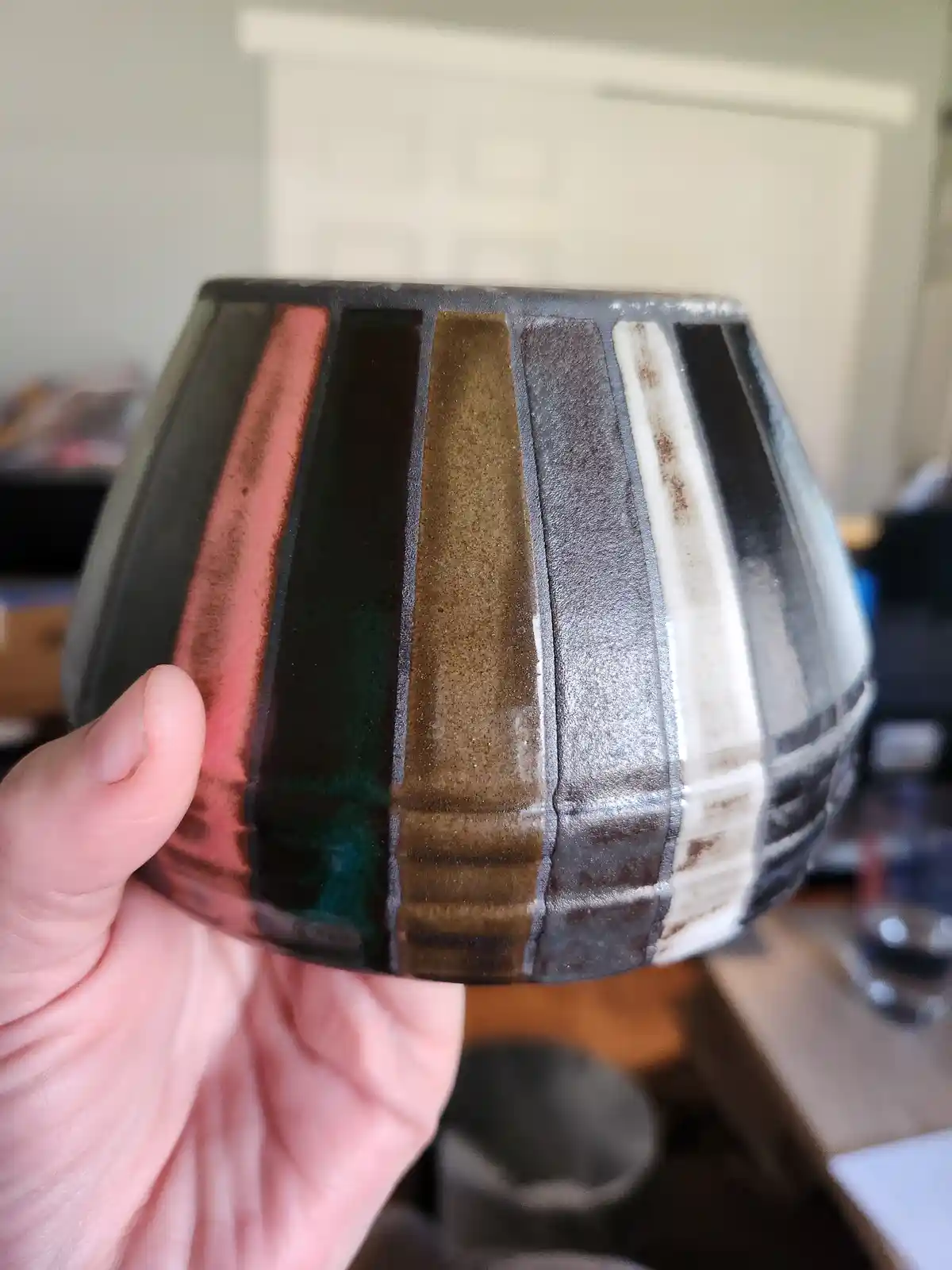 A hand holds up a ceramic bowl with vertical stripes of various colors including pink, green, brown, and white, showing off…