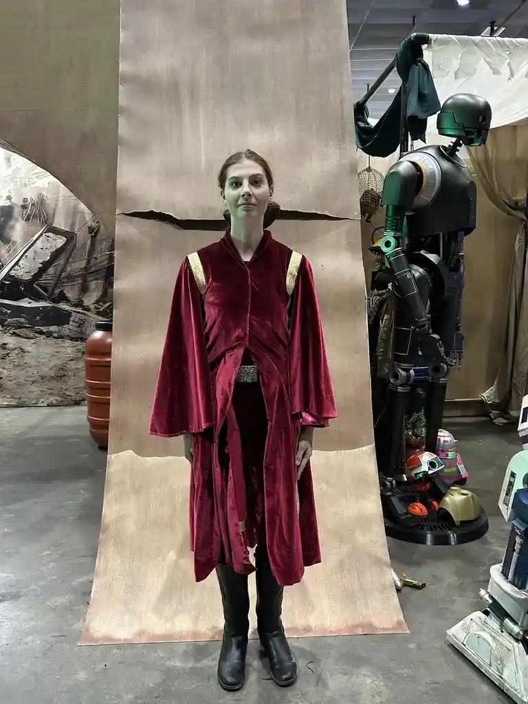 A person in a red velvet Jedi robe stands in front of a cardboard backdrop, with a green and black droid and other Star Wars…