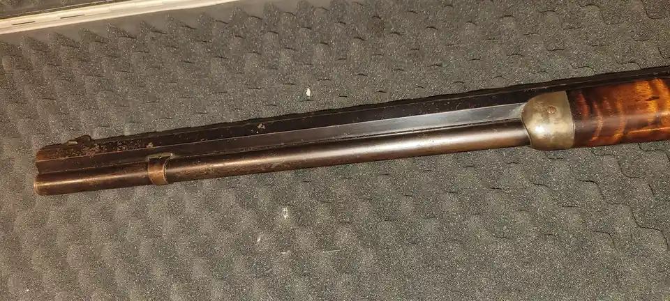 A close-up photo of an old, weathered rifle with a wooden stock and metal barrel lying on a textured gray surface. The…
