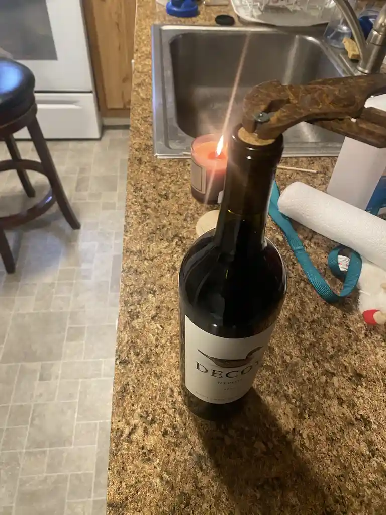 A dark wine bottle labeled 'Decoy Merlot' sits on a granite kitchen counter with a lit candle nearby. A rusty pair of pliers…