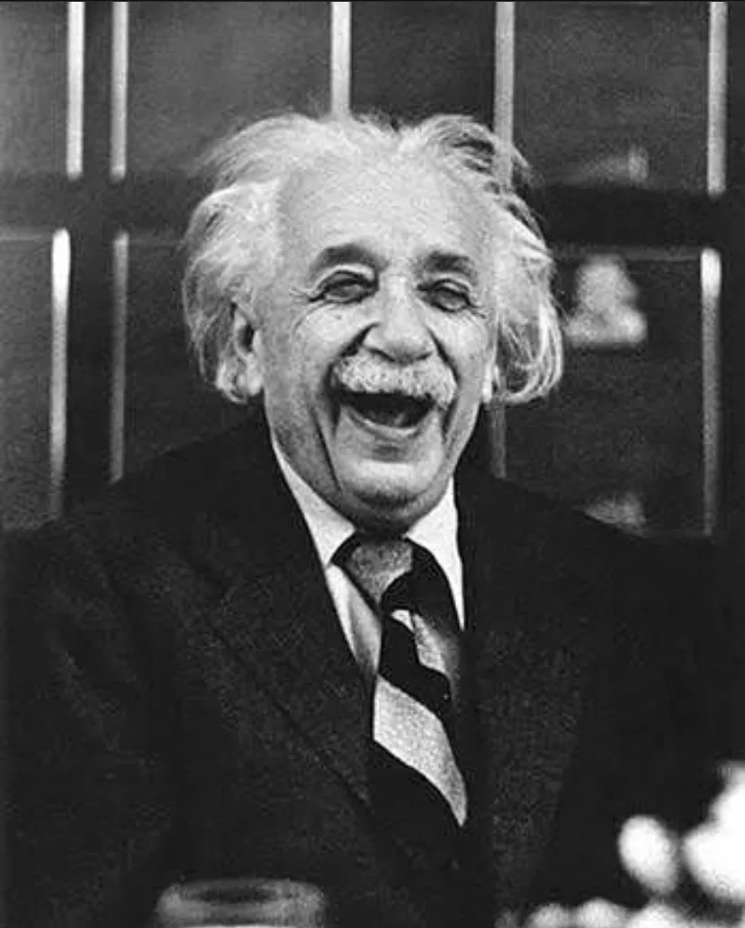 a black and white photograph of Albert Einstein laughing heartily, his eyes crinkled and mouth wide open, wearing a suit and…