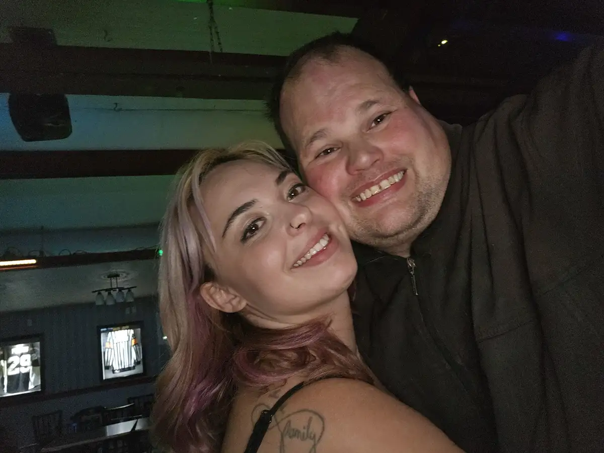 A selfie of a smiling man and woman embracing in a dimly lit bar or restaurant, with the woman's shoulder showing a tattoo…
