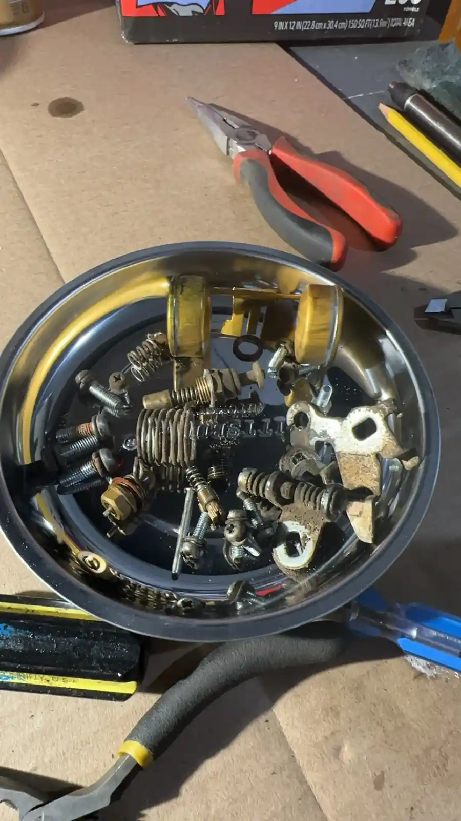 A metal bowl filled with various small, rusty car parts like springs, bolts, and brackets sits on a workbench surrounded by…