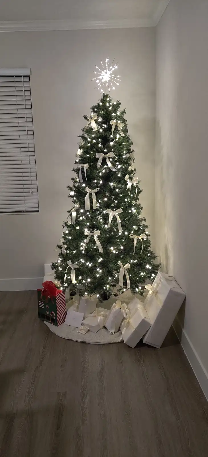 A fully decorated artificial Christmas tree with white lights and cream-colored bows stands in a corner, surrounded by…