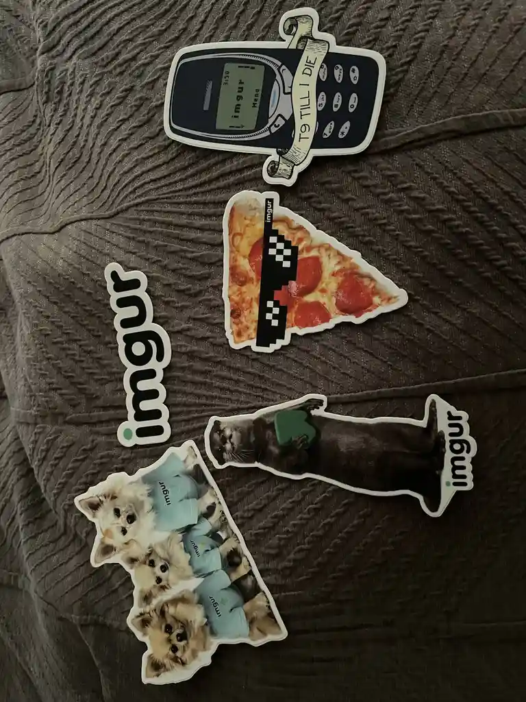 Five die-cut stickers lie on a textured gray fabric. One is a Nokia phone with 'T9 TILL I DIE' on a ribbon, another is a…