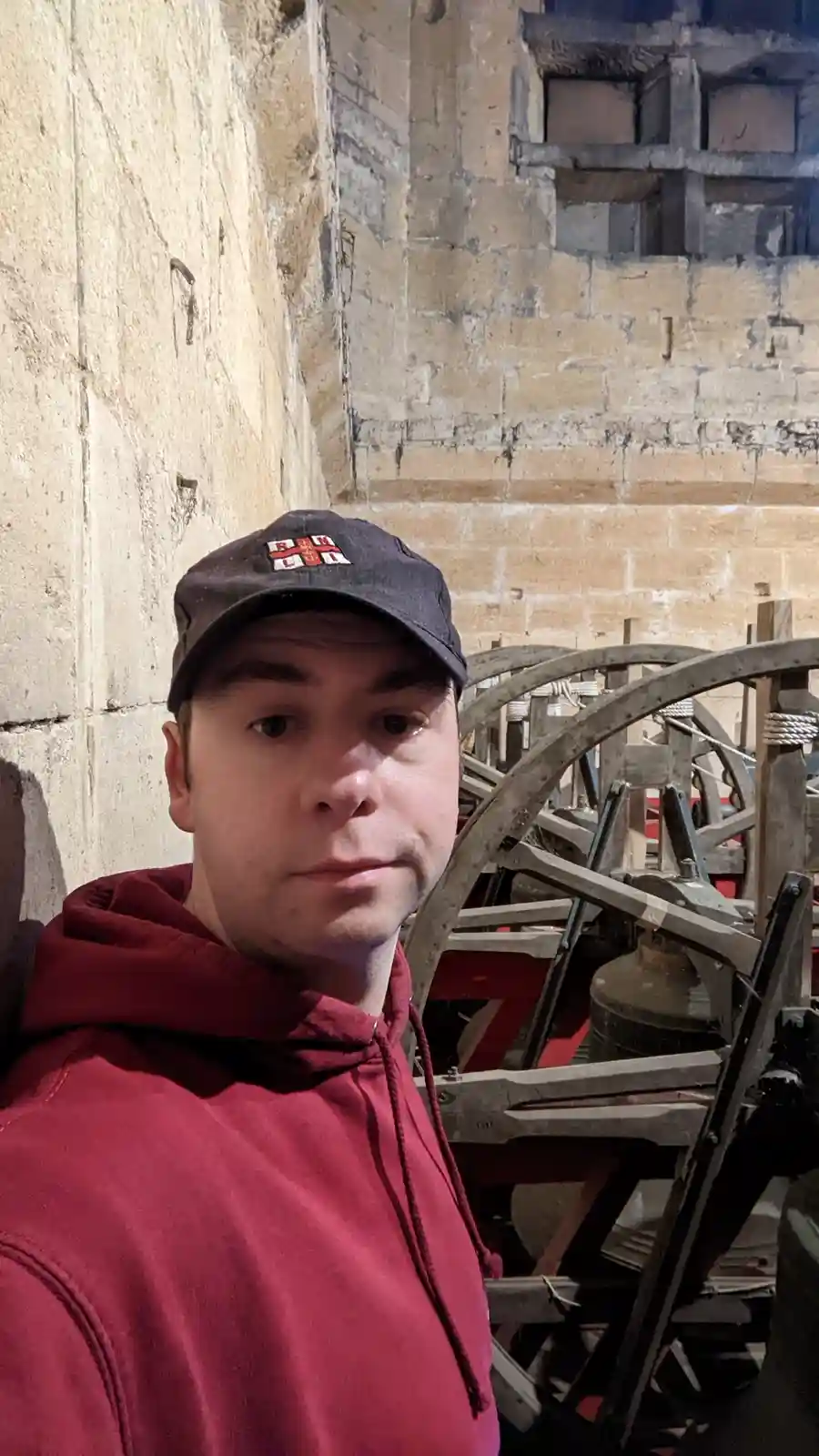 A man in a red hoodie and black cap stands in front of an old bell-ringing mechanism with large wooden wheels and a metal…