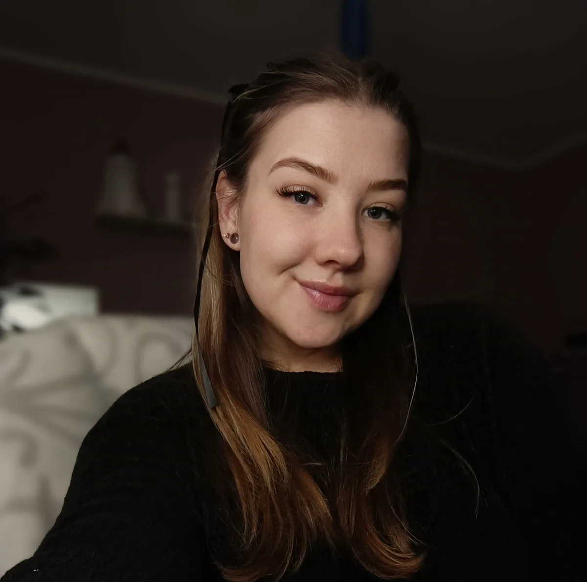 A young woman with long brown hair and a black sweater smiles subtly at the camera while wearing earphones, set against a…