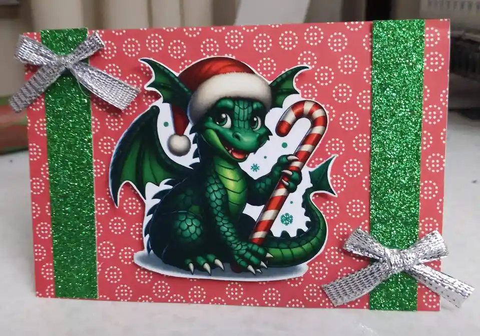 A handmade Christmas card features a cartoon green dragon wearing a red and white Santa hat and holding a red-and-white…