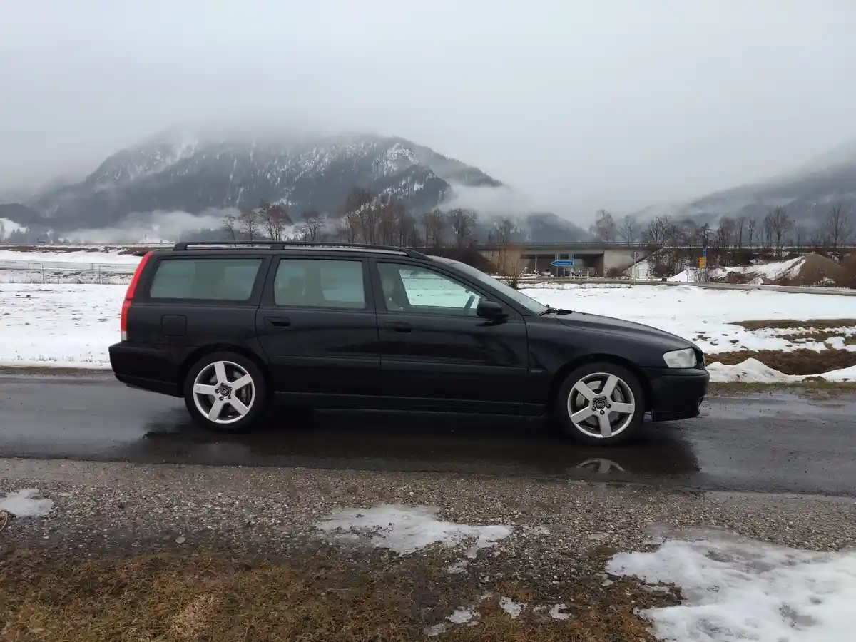 A black Volvo V70 station wagon is parked on a wet road beside melting snow and patches of grass, with fog-covered snowy…
