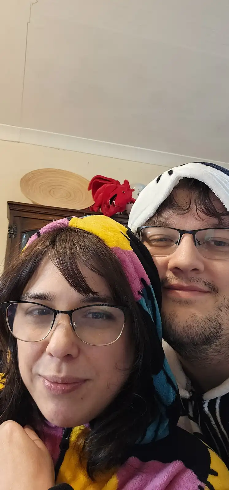 A selfie of a man and a woman in cozy, colorful hooded pajamas, both wearing glasses and smiling slightly. The woman is in…