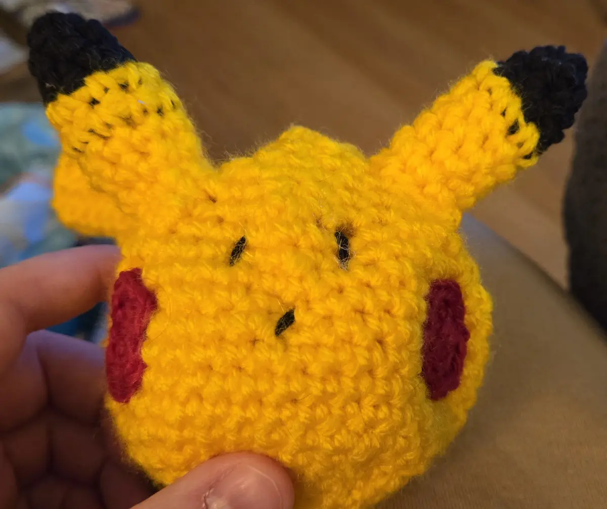A close-up photo shows a hand holding a small, crocheted Pikachu plush toy made of yellow yarn with red cheeks and…