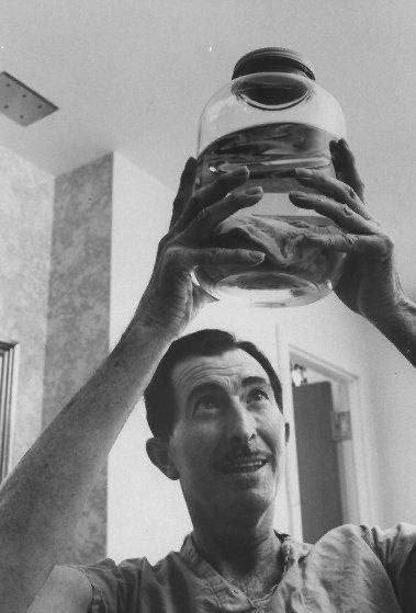 A black-and-white photo shows a man holding a large glass jar above his head, looking upward with a slight smile. The jar…