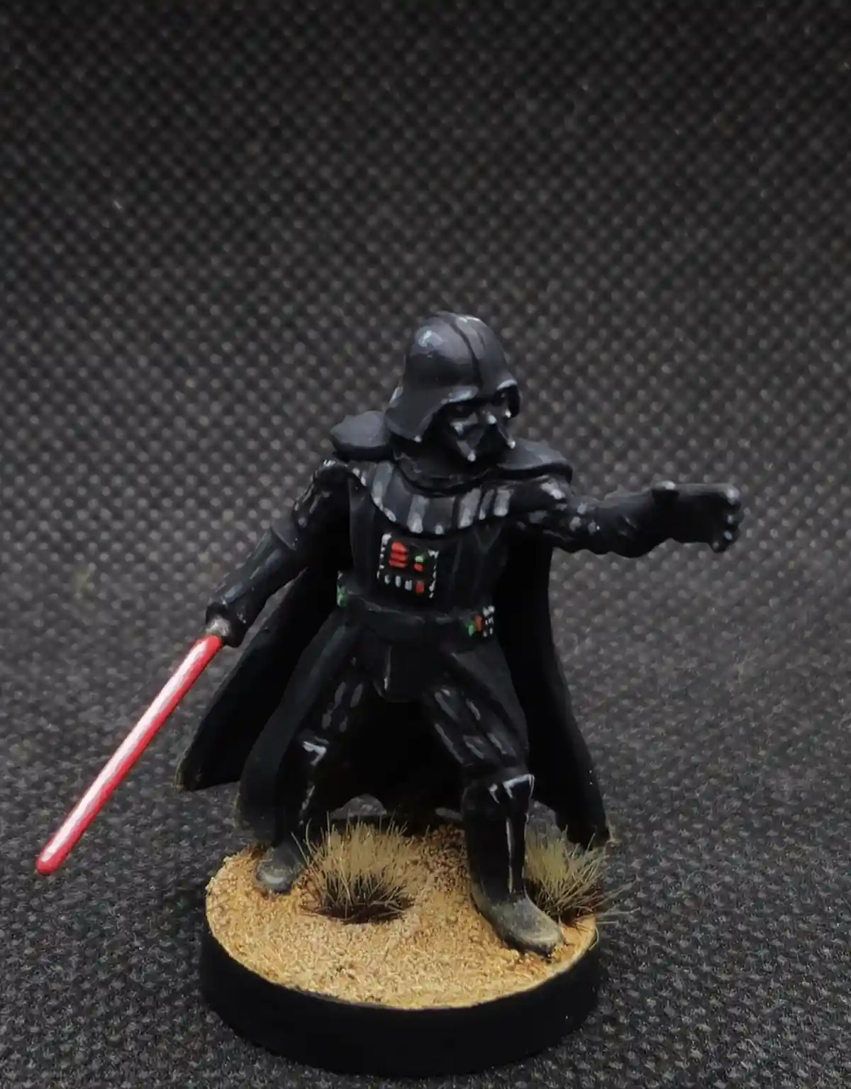 A painted Darth Vader miniatures figure stands on a sandy base with sparse grass, holding a red lightsaber in one hand and…