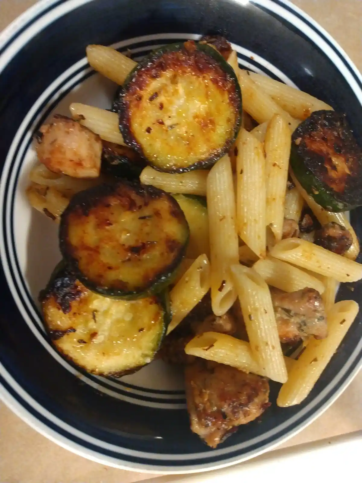 A close-up photo shows a bowl of penne pasta mixed with charred zucchini slices and pieces of cooked sausage. The bowl has a…