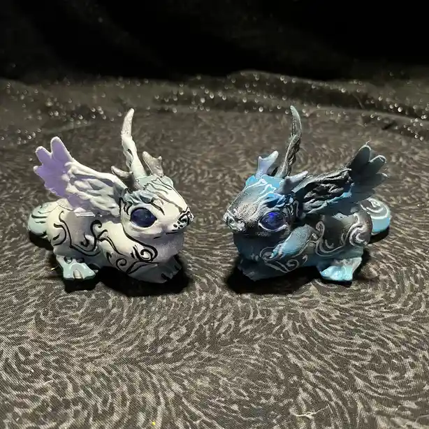 Two small, stylized dragon figurines with wings and horns sit side by side on a dark, textured fabric. The dragon on the…