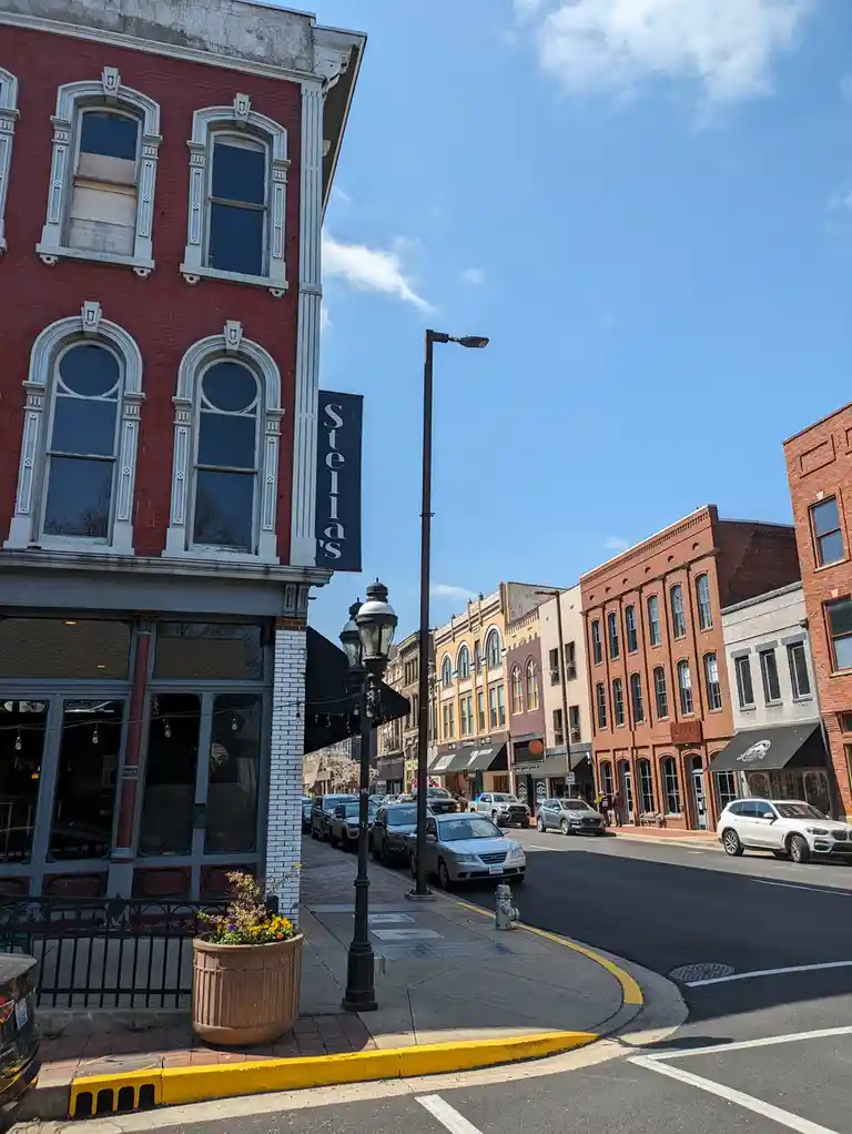A sunny street view of a historic downtown area with red brick buildings, parked cars, and a prominent black vertical sign…