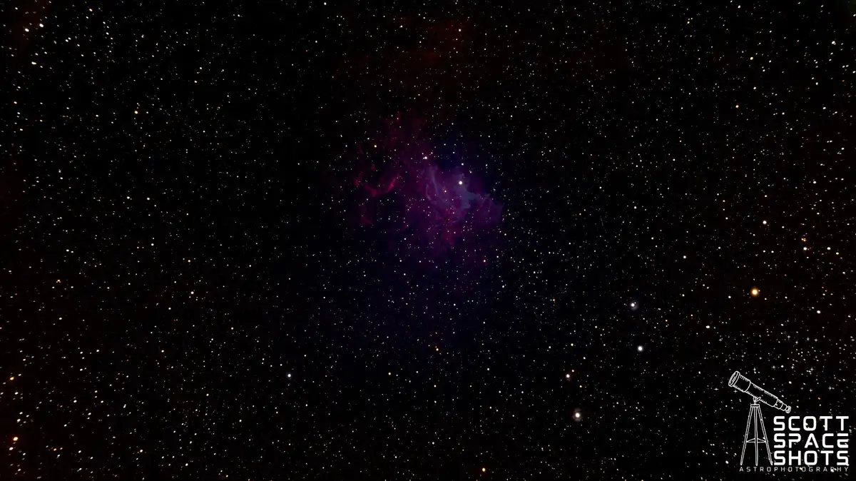 A deep-sky astrophotograph shows a dark starfield with a prominent purple and red nebula at center, surrounded by countless…