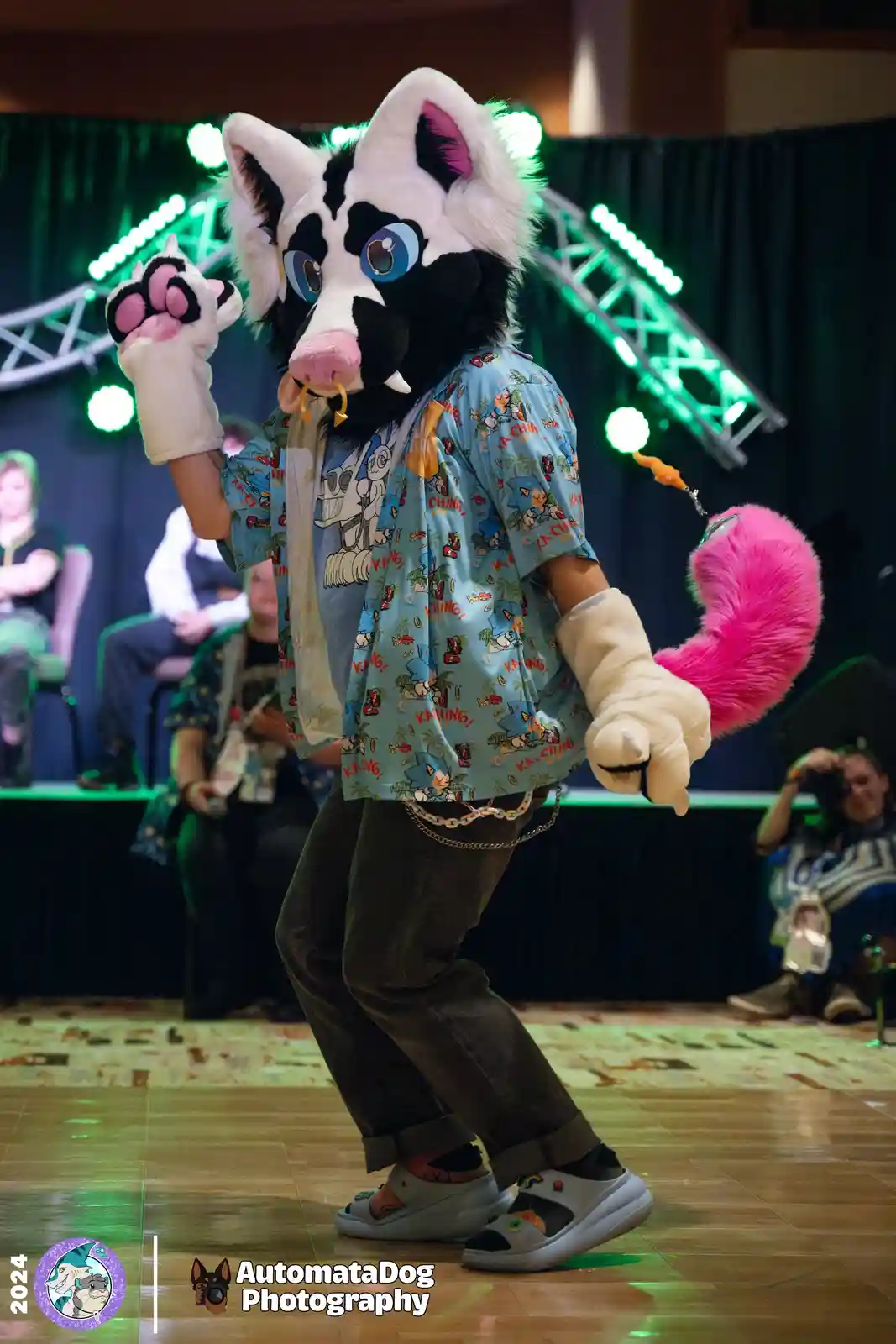 A person in a black-and-white fursuit with large blue eyes, pink nose, and a bright pink tail is posing on a stage with…
