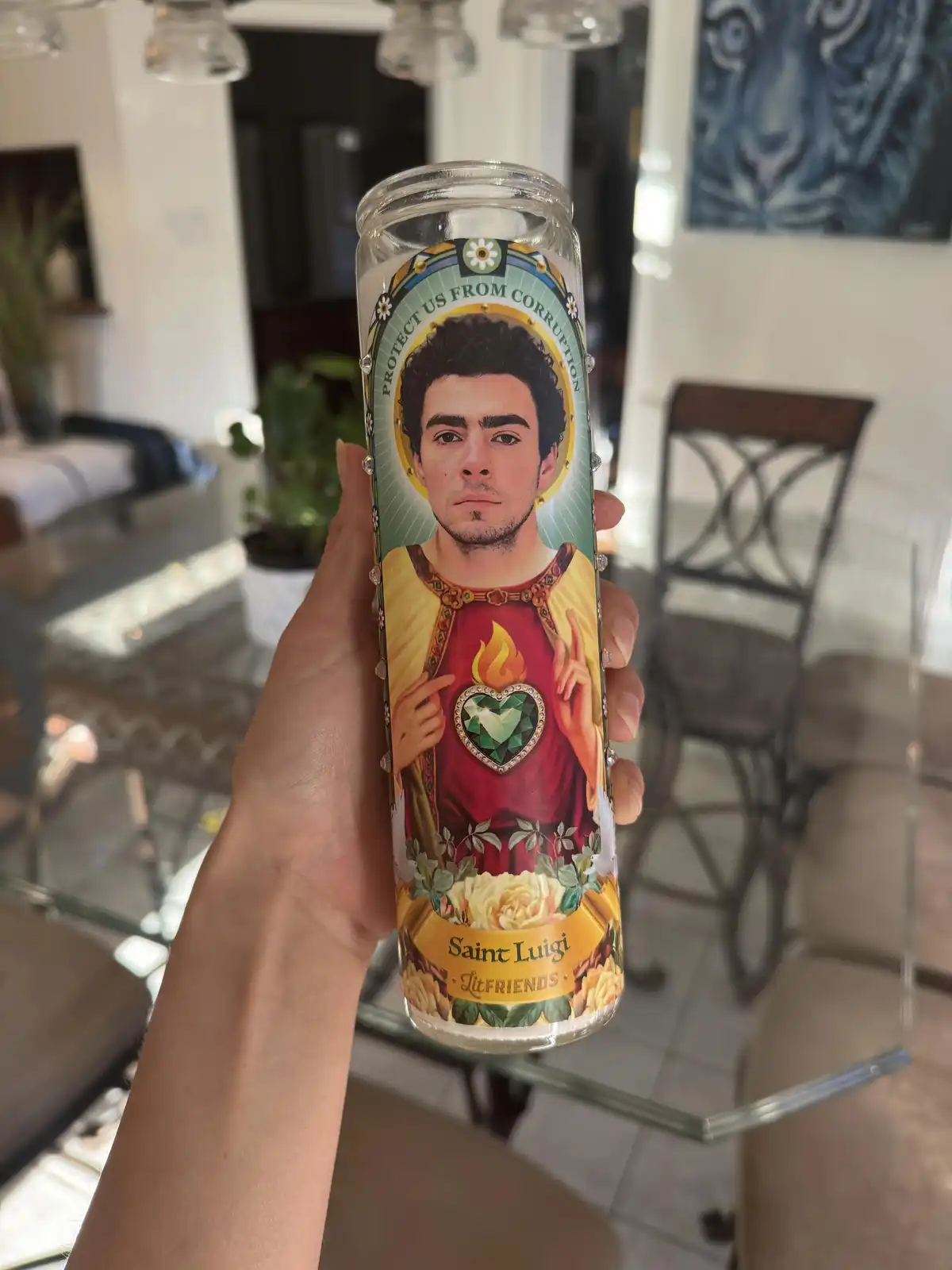 A hand holds a tall, clear glass prayer candle with a printed label depicting a man with dark hair and a goatee, dressed in…