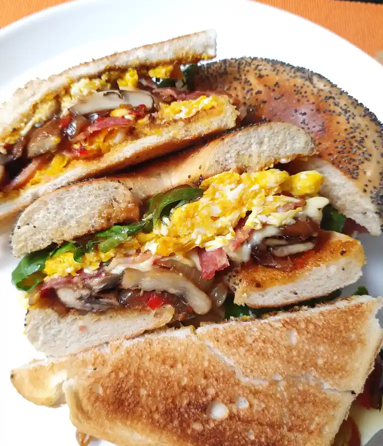 A close-up photo shows a bagel sandwich cut in half, revealing a filling of scrambled eggs, bacon, mushrooms, melted cheese…